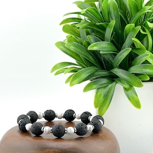 May include: A bracelet made of black lava stone and white gemstone beads. The bracelet has large, textured black beads and smaller, round white beads. It is displayed on a wooden surface, with a green plant in the background.