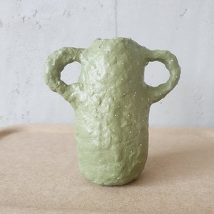 Könnte beinhalten: Eine hellgrüne Keramikvase mit zwei Henkeln und einer strukturierten Oberfläche. Die Vase ist mit weißen Punkten verziert.