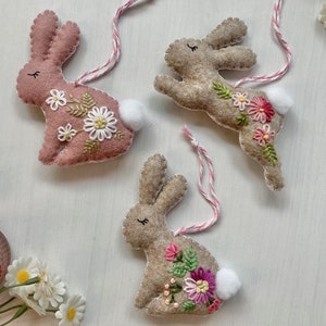 May include: Three felt bunny ornaments with embroidered flowers and white pom-poms. The bunnies are pink, brown, and beige.