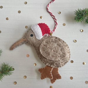 May include: A handmade felt kiwi bird Christmas decoration. The bird is mainly tan with brown feet and a long beak. It wears a red and white Santa hat. A red and white string is attached for hanging.