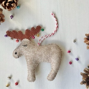 May include: A felt moose Christmas ornament with brown antlers decorated with colourful bells. The moose is light brown with closed eyes and a red and white string for hanging. Pine cones and small decorative beads surround the ornament.