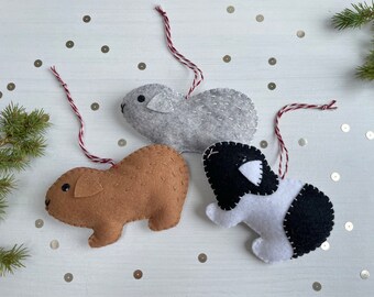 May include: Three felt ornaments shaped like guinea pigs. The guinea pigs are brown, gray, and black and white. Each ornament has a red and white string attached for hanging.