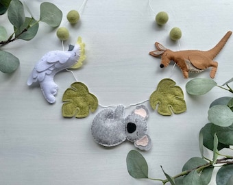 May include: A felt garland with a koala, a cockatoo, and a kangaroo, each hanging from a green felt leaf. The garland is strung with white thread and green felt balls.