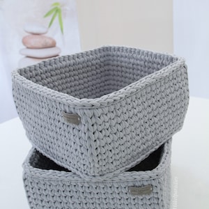 Op de afbeelding: Twee grijze gehaakte opbergmanden die op elkaar gestapeld zijn. De manden zijn gemaakt met een textuur-steek en hebben een klein label aan de voorkant met de tekst "Handmade with Love".