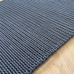 Könnte beinhalten: Ein dunkelgrauer Strickteppich mit einer dicken, strukturierten Webart. Der Teppich ist aus einem weichen, gemütlichen Material gefertigt und hat ein schlichtes, modernes Design.