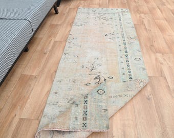 Puede incluir: Una alfombra vintage de color beige claro y azul con un patrón floral y un borde negro. La alfombra está doblada por la mitad y se encuentra sobre un suelo de madera.