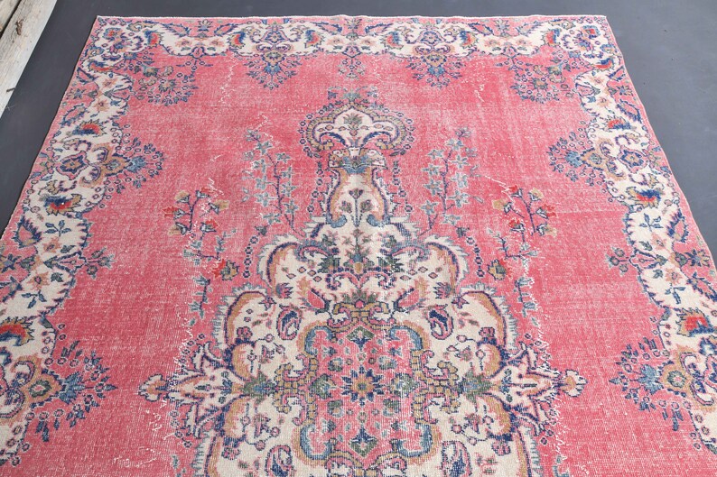 K&ouml;nnte beinhalten: Ein vintagefarbener rosa Teppich mit einem floralen Muster. Der Teppich hat ein zentrales Medaillon mit komplizierten Details und einen Rand mit einem sich wiederholenden floralen Muster.