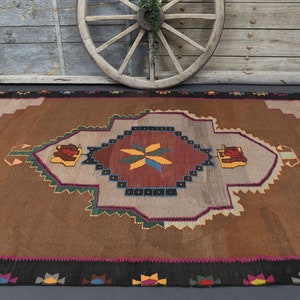 May include: A brown and beige rug with a geometric pattern in shades of red, green, blue, and yellow. The rug has a central medallion with a starburst design.