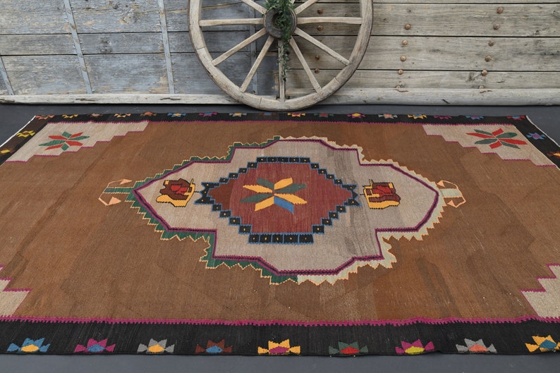 May include: A brown and beige rug with a geometric pattern in shades of red, green, blue, and yellow. The rug has a central medallion with a starburst design.