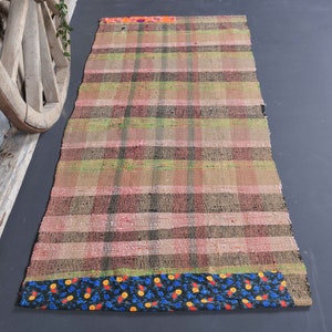 May include: A rectangular, handwoven rug with a plaid pattern in shades of pink, green, and brown. The rug has a decorative border with a blue floral design. The rug is placed on a dark gray surface.