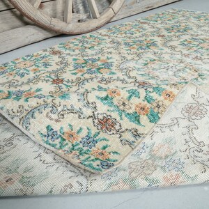 May include: A vintage rug with a floral pattern in shades of blue, green, and orange on a cream background. The rug is folded in half, showing the back side, which is a lighter cream color with a subtle pattern.