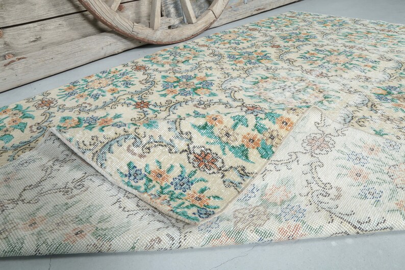 May include: A vintage rug with a floral pattern in shades of blue, green, and orange on a cream background. The rug is folded in half, showing the back side, which is a lighter cream color with a subtle pattern.