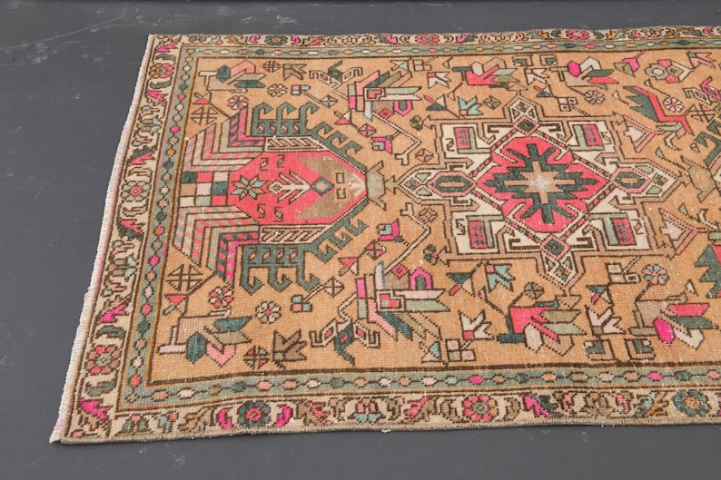 Puede incluir: Una alfombra turca vintage con fondo beige y intrincados patrones geom&eacute;tricos en tonos rosa, verde y marr&oacute;n. La alfombra tiene un borde con flecos.