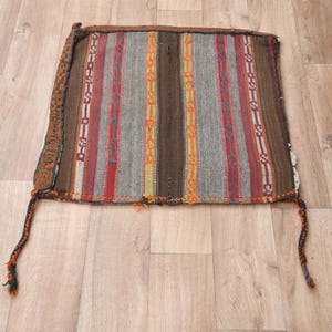 May include: A rectangular, woven textile with vertical stripes in shades of brown, red, gray, and yellow. The edges are finished with a darker brown border and tassels. The textile is lying on a light wood surface.