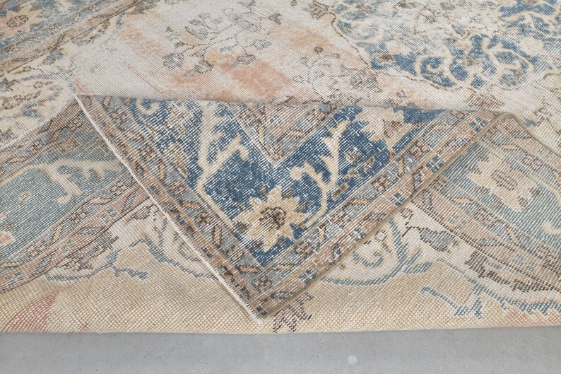 May include: Close-up of a beige rug with a blue and brown floral pattern. The rug is folded in half, showing the underside of the fabric.