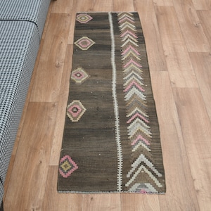 May include: A long, narrow rug with a brown background and a colorful geometric pattern. The pattern features pink, white, and brown triangles and diamonds.