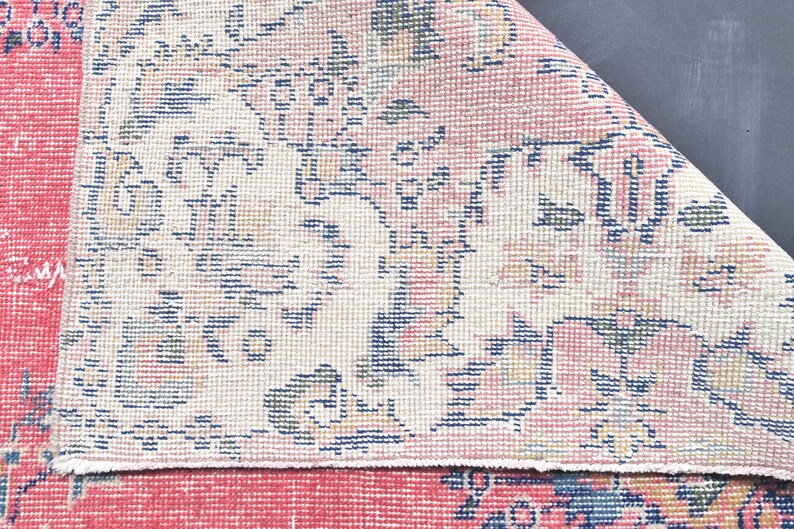 K&ouml;nnte beinhalten: Nahaufnahme eines rosa-wei&szlig;en Teppichs mit Blumenmuster. Der Teppich hat einen Fransenrand und ist aus einem weichen, gewebten Material gefertigt.