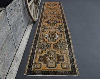 May include: A long, narrow rug with a geometric pattern in shades of brown, beige, and black. The rug has a fringe on both ends.