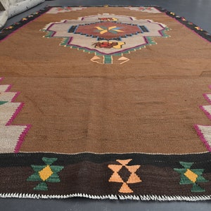 May include: A brown, handwoven rug with a geometric pattern in shades of red, green, yellow, and orange. The rug has a fringed border.