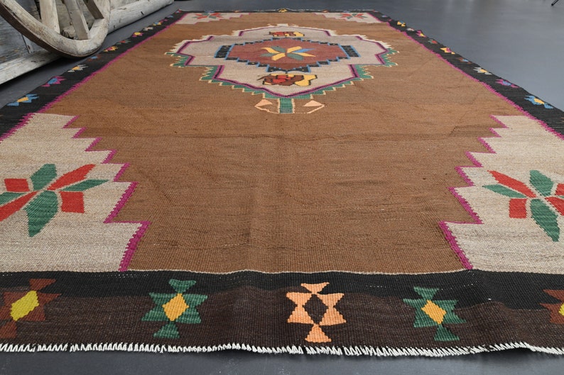 May include: A brown, handwoven rug with a geometric pattern in shades of red, green, yellow, and orange. The rug has a fringed border.