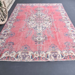 K&ouml;nnte beinhalten: Ein vintagefarbener rosa Teppich mit einem floralen Muster und einem wei&szlig;en Rand. Der Teppich hat einen abgenutzten Look und ist aus Wolle.