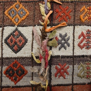 May include: A close-up of a woven rug with a geometric pattern in shades of red, orange, brown, and gray. The rug has a fringe border and is decorated with colorful fabric scraps.