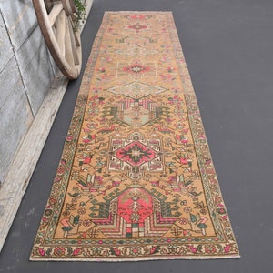 Puede incluir: Una alfombra larga y rectangular con un dise&ntilde;o persa tradicional. La alfombra es principalmente beige con acentos de rosa, verde y negro. El dise&ntilde;o presenta intrincados patrones florales y geom&eacute;tricos.
