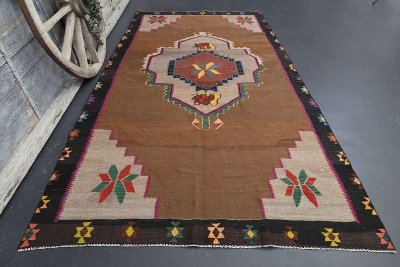 May include: A brown and beige rug with a colourful geometric pattern. The rug has a central medallion with a starburst design and a border with repeating diamond shapes.
