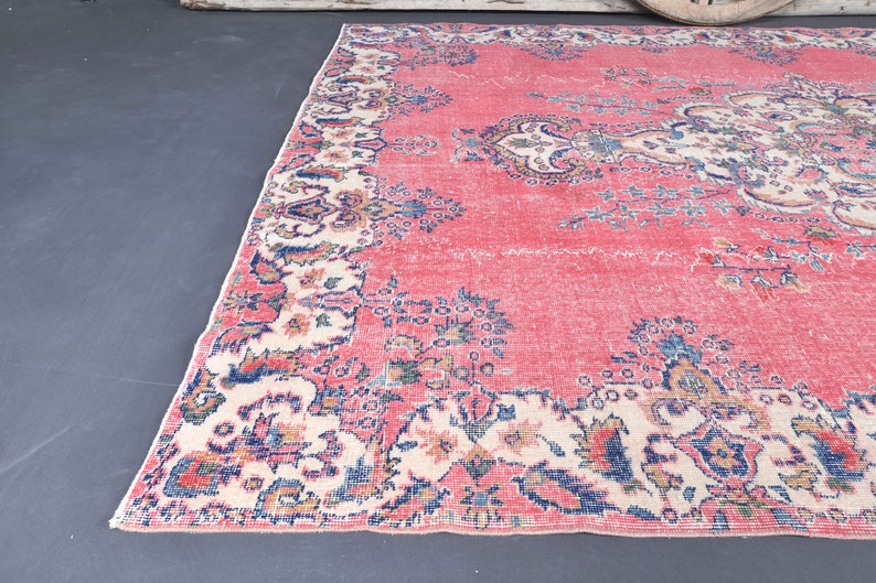 K&ouml;nnte beinhalten: Ein Vintage-Teppich mit rosafarbenem Hintergrund und einem floralen Muster in Blaut&ouml;nen, Creme und Braun. Der Teppich hat einen Fransenrand.