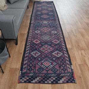 May include: A long, narrow runner rug featuring a repeating geometric diamond pattern in shades of deep purple, navy blue, and soft pink. The rug has a dark border with a checkered design at the ends and a textured, woven appearance throughout.