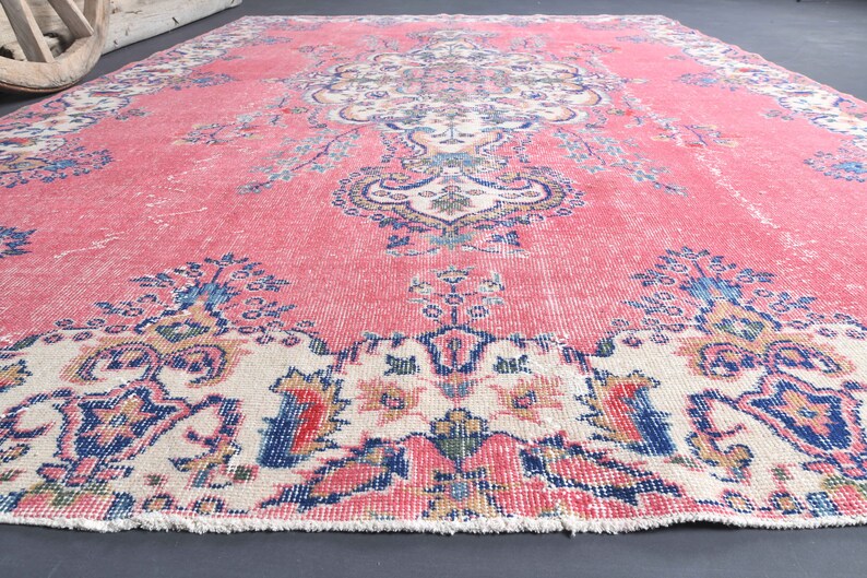 K&ouml;nnte beinhalten: Ein vintagefarbener rosa und cremefarbener Teppich mit einem Blumenmuster. Der Teppich hat einen Fransenrand und ist aus Wolle.