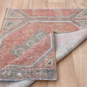 May include: A rectangular, distressed rug with a geometric design in shades of red, gray, and beige. The rug's corner is folded back, revealing the underside. The rug is placed on a light-colored wooden floor.