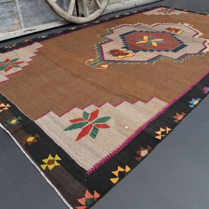 May include: A brown and beige rug with a geometric pattern in shades of red, green, yellow, and blue. The rug has a black border with a pink accent.