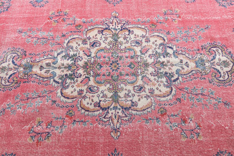 K&ouml;nnte beinhalten: Ein Vintage-Teppich mit rosafarbenem Hintergrund und einem detaillierten floralen Muster in Blau-, Wei&szlig;- und Beiget&ouml;nen. Der Teppich hat einen abgenutzten und verblassten Look, der ihm einen einzigartigen Charakter verleiht.