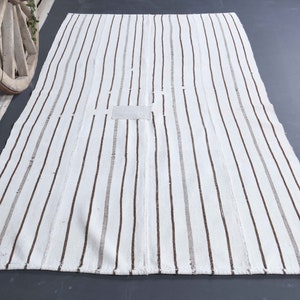 Puede incluir: Una alfombra rectangular de color blanquecino con rayas verticales marrones. La alfombra tiene un diseño sencillo y una paleta de colores neutros. La alfombra está hecha de un material tejido y tiene un parche rectangular en el centro.