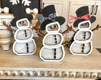 May include: Three snowman-shaped shaker ornaments with black top hats, orange carrot noses, and brown stick arms. Each snowman has a clear center filled with small white beads and black button accents. One snowman has a black and white gingham bow.