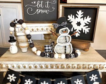May include: A wooden tiered tray display featuring a black sign with white text reading let it snow, a black sign with a white snowflake, a snowman figure with clear sections filled with beads, and a garland of black, white, and plaid wooden beads.