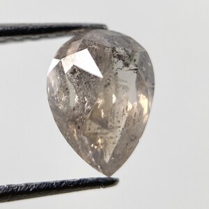 May include: A pear-shaped diamond, held by black tweezers, with a light brown colour and visible inclusions. The faceted gem is displayed against a white background, showcasing its polished surface and unique shape.