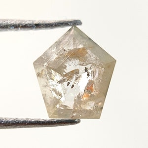 May include: A clear, pentagon-shaped diamond held by tweezers. The gemstone has a light brown hue with internal inclusions and facets. The diamond is displayed against a white background, highlighting its clarity and shape.