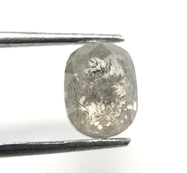 May include: A faceted, oval-shaped, gray diamond held by metal tweezers. The diamond has internal inclusions and a polished surface, reflecting light. The gemstone is set against a white background.
