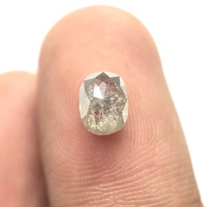 May include: A close-up of a light gray, oval-shaped diamond with a faceted surface. The diamond has internal inclusions and is held against a white background.