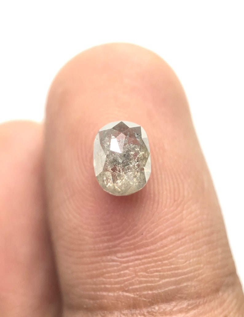 May include: A close-up of a light gray, oval-shaped diamond with a faceted surface. The diamond has internal inclusions and is held against a white background.