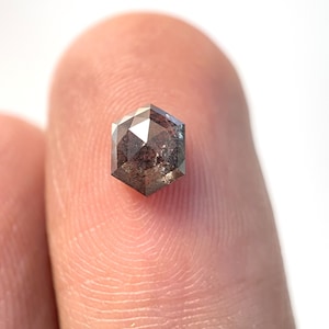 Op de afbeelding: Een gefacetteerde, zeshoekig geslepen diamant met een donkergrijze en bruine kleur. De edelsteen wordt tussen twee vingers gehouden, wat de unieke vorm en reflecterende oppervlakken benadrukt. De facetten van de diamant vangen het licht op en creëren een sprankelend effect.
