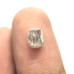 May include: A faceted, oval-shaped diamond with a salt and pepper appearance. The gemstone has a mix of gray and clear tones, with a hint of yellow. The diamond is held between two fingers against a white background.