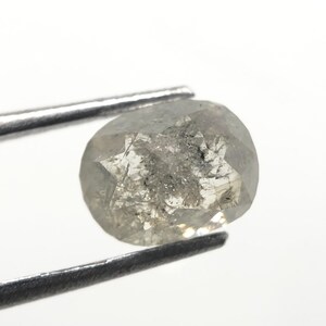 May include: A faceted, oval-shaped, gray diamond held by metal tweezers. The diamond has internal inclusions and a star-like pattern on its surface. The background is plain white.