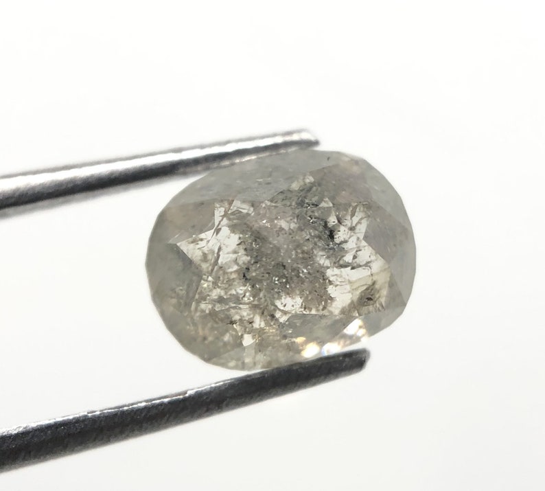 May include: A faceted, oval-shaped, gray diamond held by metal tweezers. The diamond has internal inclusions and a star-like pattern on its surface. The background is plain white.