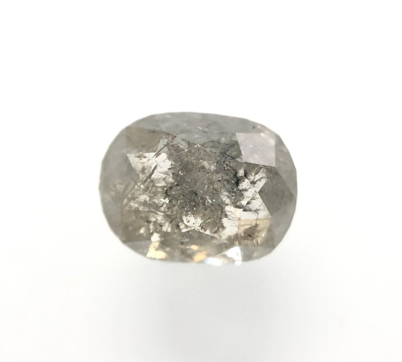 May include: A faceted, oval-shaped, salt and pepper diamond. The gemstone has a mix of gray and white tones with internal inclusions. The diamond is set against a white background, showcasing its natural beauty.