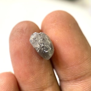 May include: A small, rough-cut, grey gemstone with a textured surface. The stone is held between two fingers, showcasing its natural, unpolished appearance. The gem has a slightly elongated shape and a subtle sparkle.