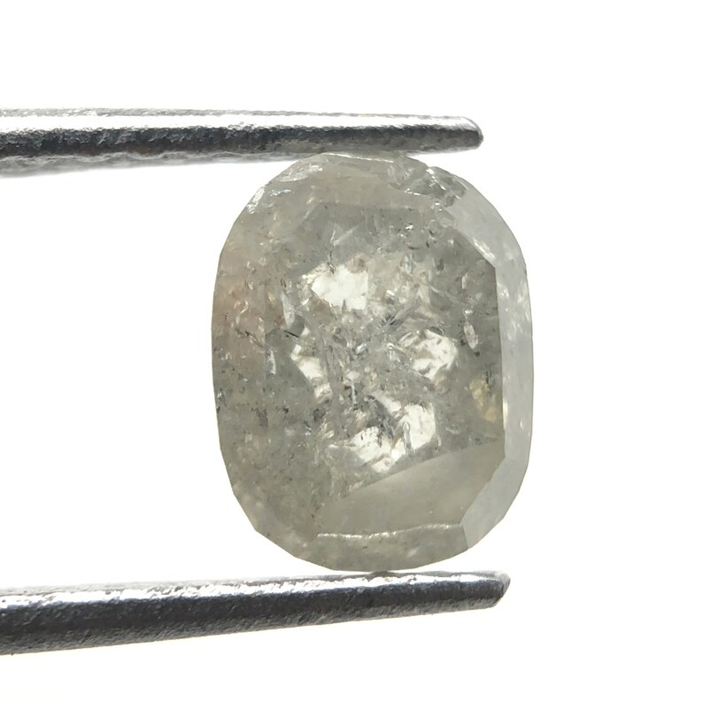May include: A faceted, oval-shaped, light gray diamond held by tweezers. The diamond has internal inclusions and a slightly irregular surface, reflecting light. The gem is set against a white background.