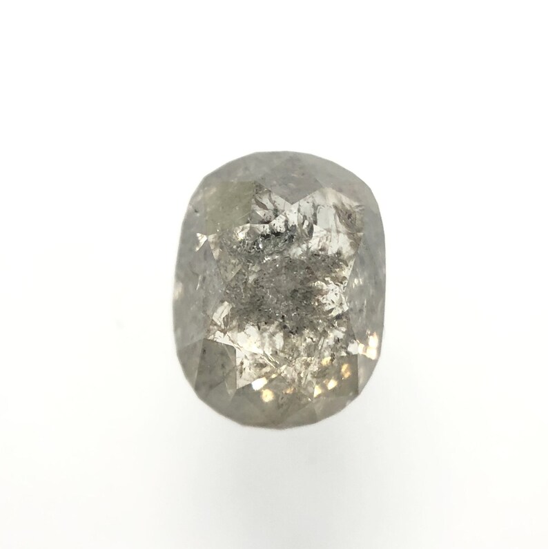May include: A faceted, oval-shaped, salt and pepper diamond. The gemstone has a gray hue with dark inclusions and a brilliant, reflective surface. The diamond is set against a white background.